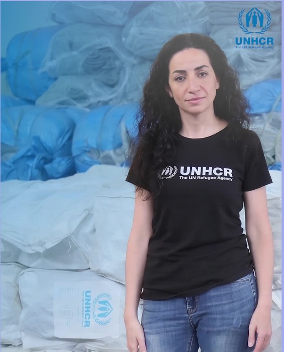 UNHCR field video frame with a staff member standing in front of stacked relief supplies